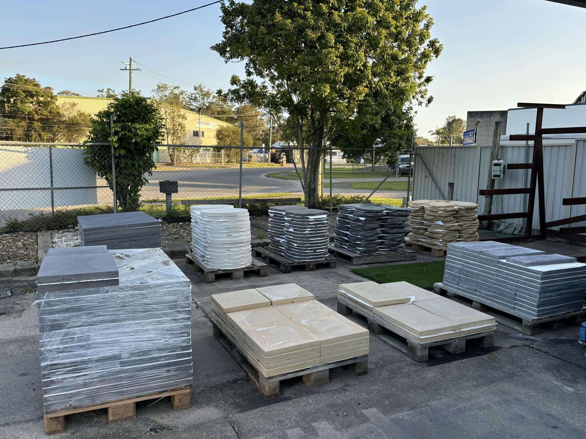 463753108_998338912306409_3981364455454672842_n Outdoor storage area with stacked flat panels in white, beige, and gray, neatly arranged on pallets, wrapped in plastic, surrounded by fencing and trees.